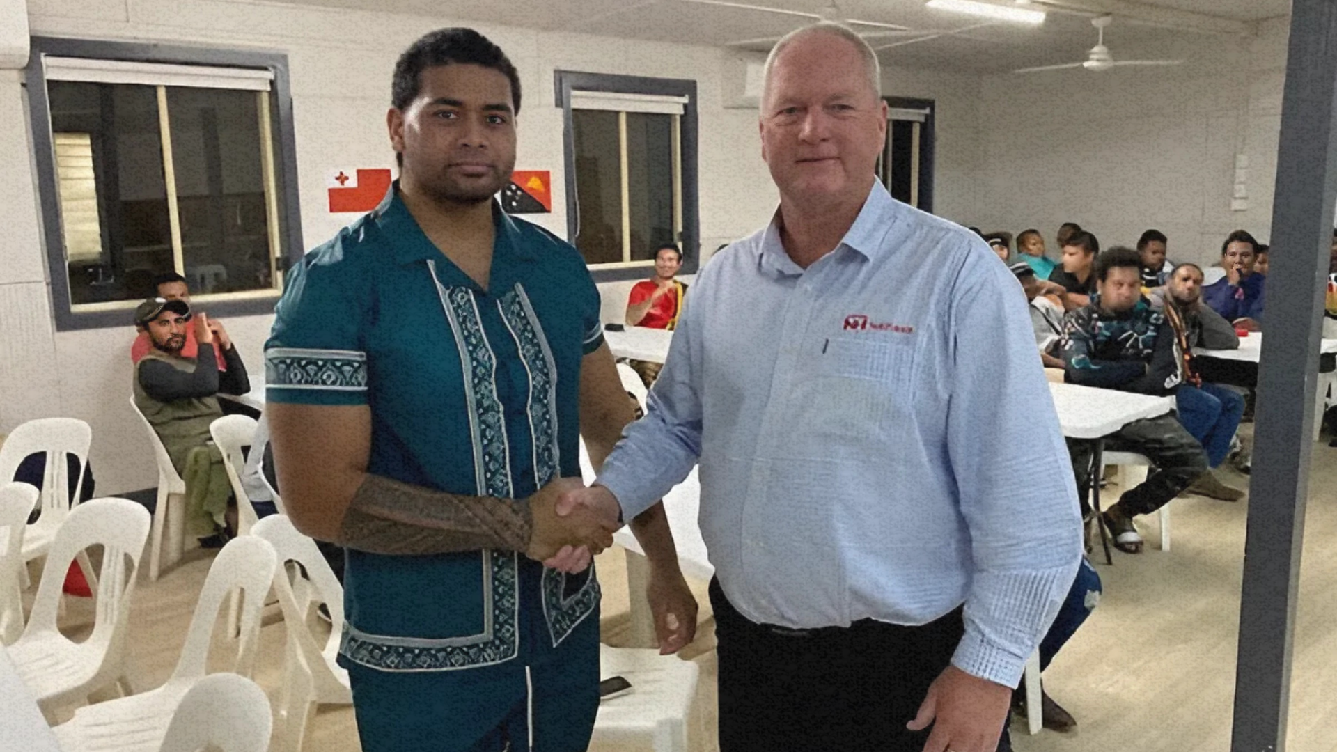 Two men are shaking hands in a room with tables and chairs.