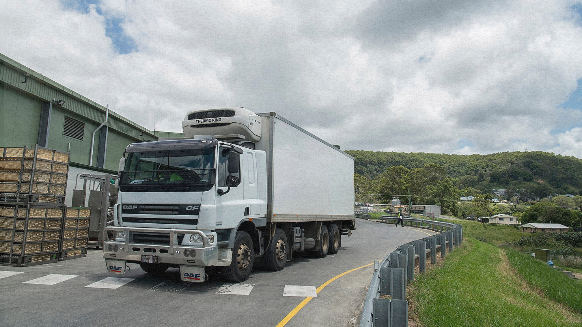 A refrigerated truck parked at a logistics facility, with crates nearby and a winding road leading i