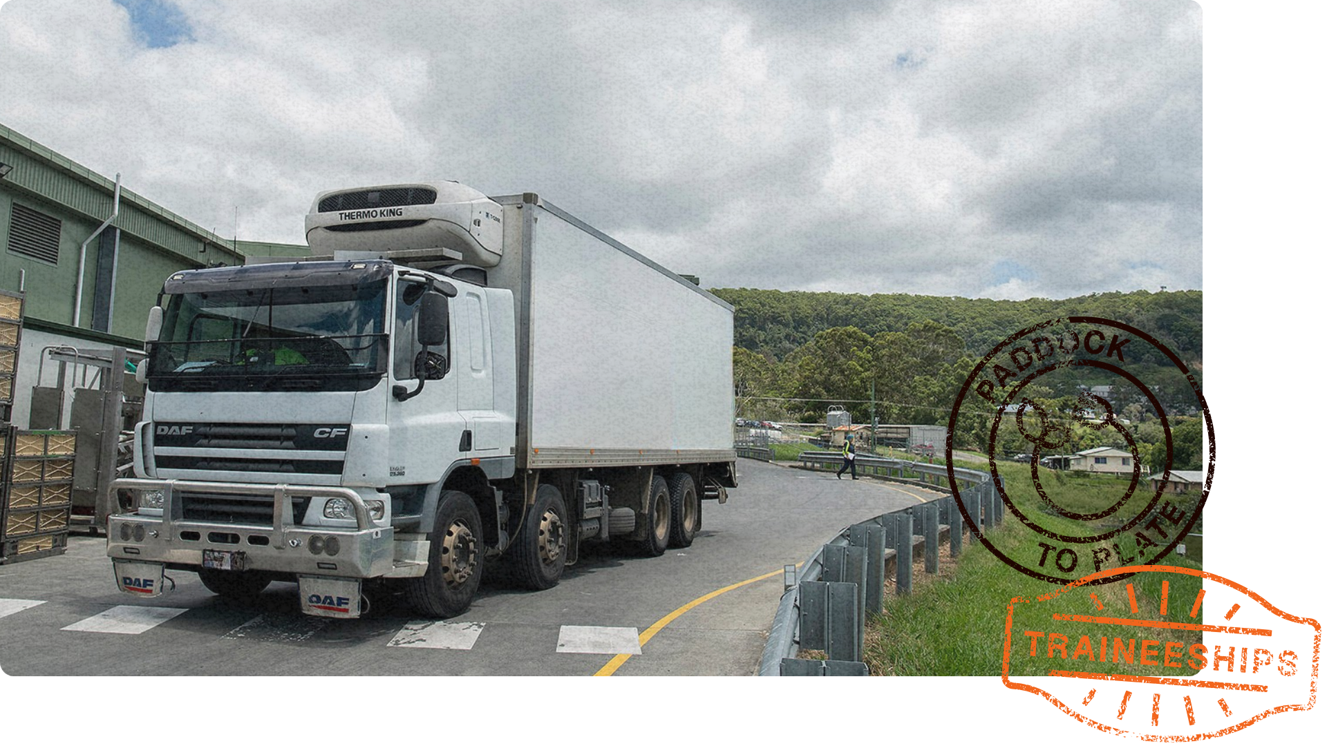 A refrigerated truck parked at a logistics facility, with crates nearby and a winding road leading into a hilly, rural area.