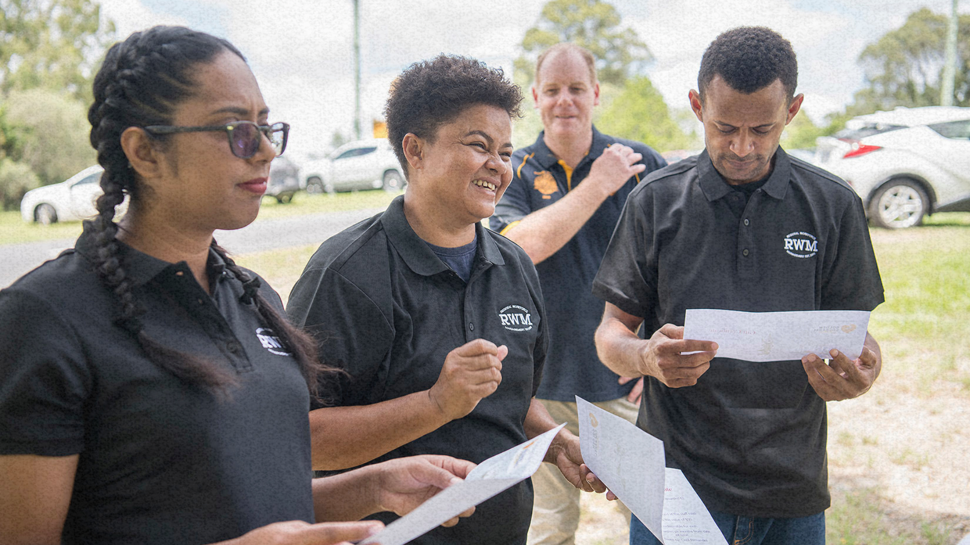 A group of workers in black RWM shirts review certificates outdoors, with one smiling and engaging.