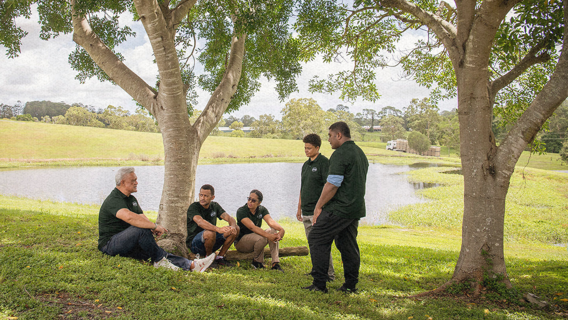 Five PALM scheme workers in green shirts sit and stand under a tree by a pond, engaged in conversati