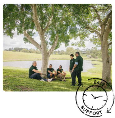Five PALM scheme workers in green shirts sit and stand under a tree by a pond, engaged in conversation. 'Approved PALM Employer' stamp in the corner.