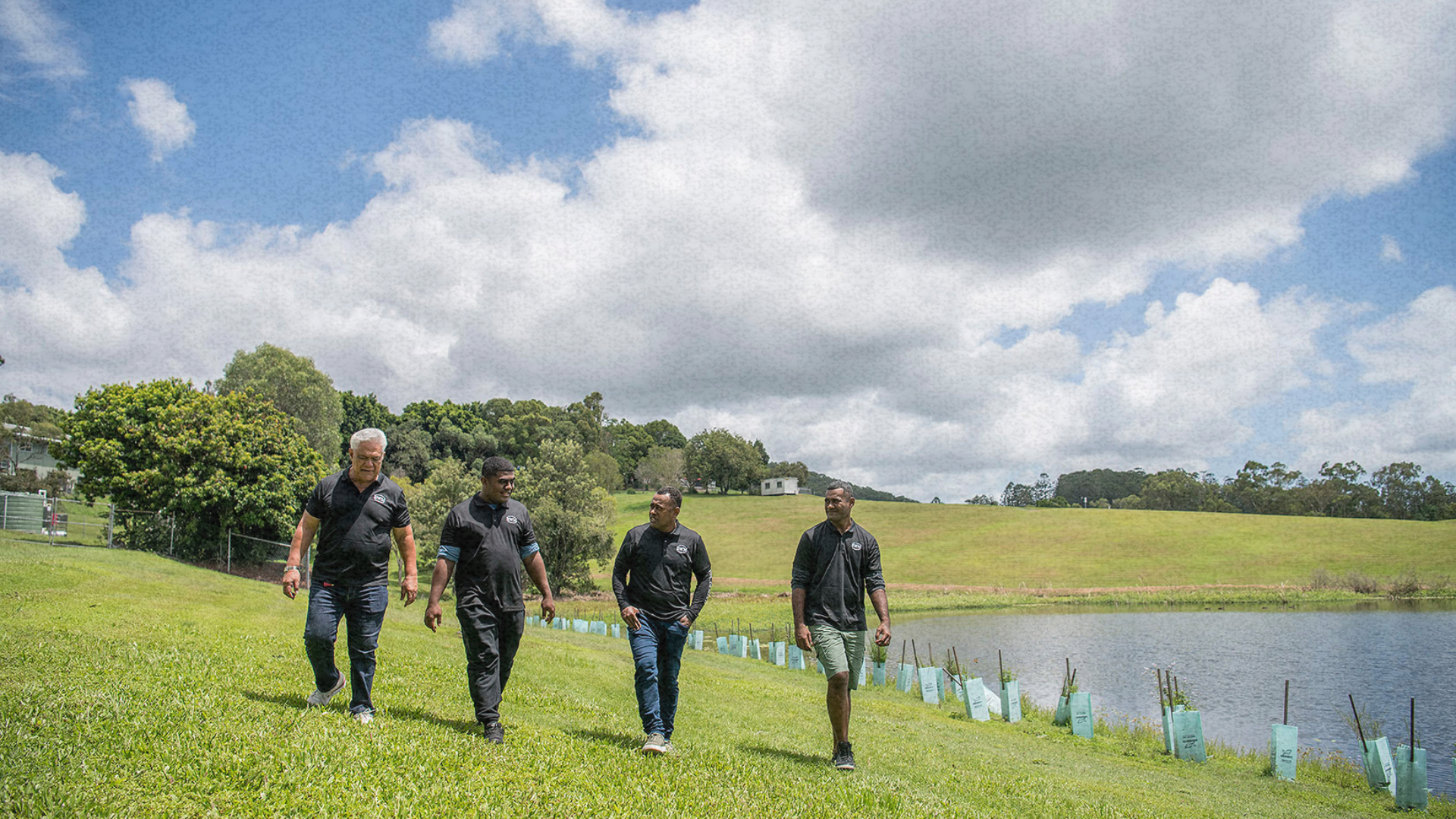 Four men in green RWM shirts walk along a grassy hillside near a pond, surrounded by rolling green f