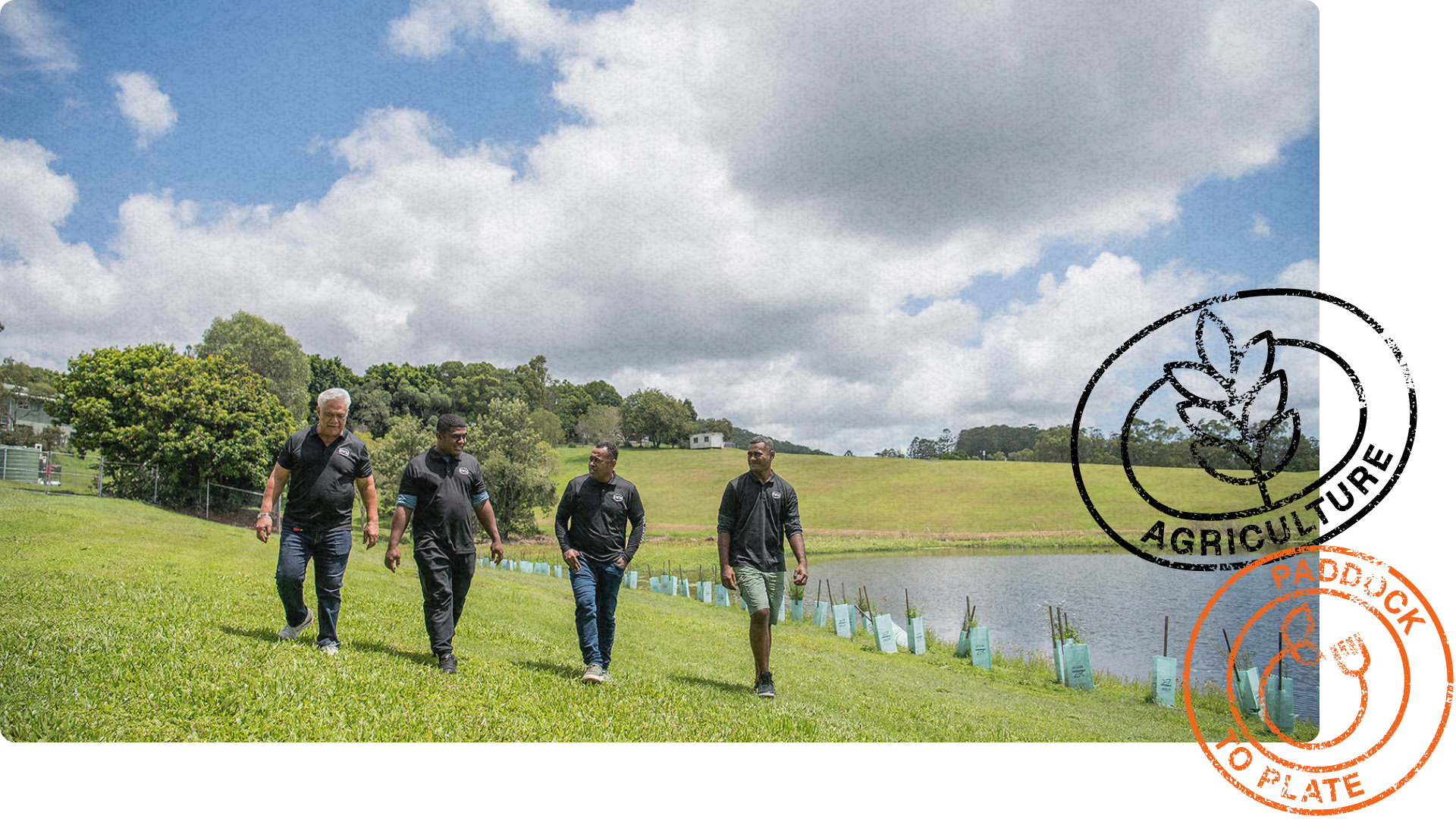Four men in green RWM shirts walk along a grassy hillside near a pond, surrounded by rolling green fields and young tree plantings.