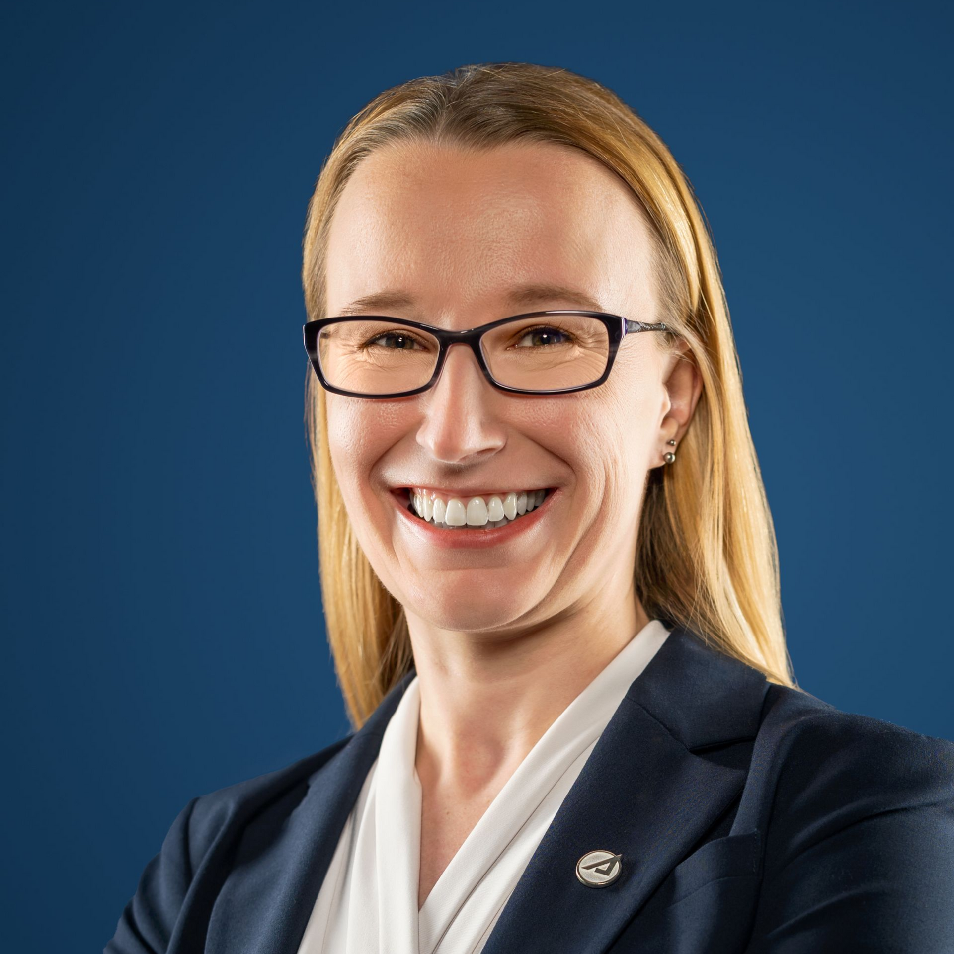Woman smiling, wearing glasses, outdoors in front of blurred trees.