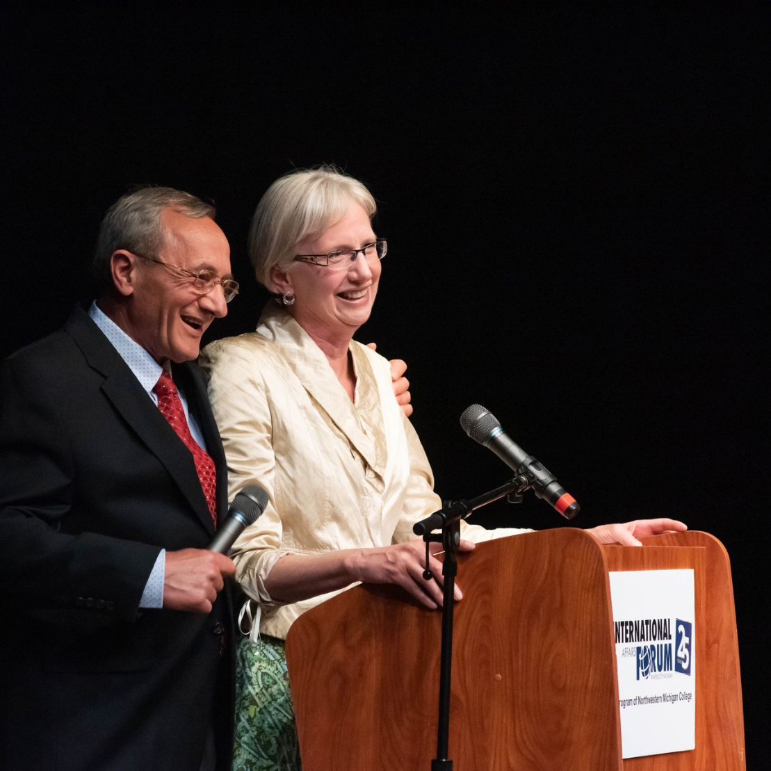 Two speakers smiling at podiums on a dark stage, each with a microphone.