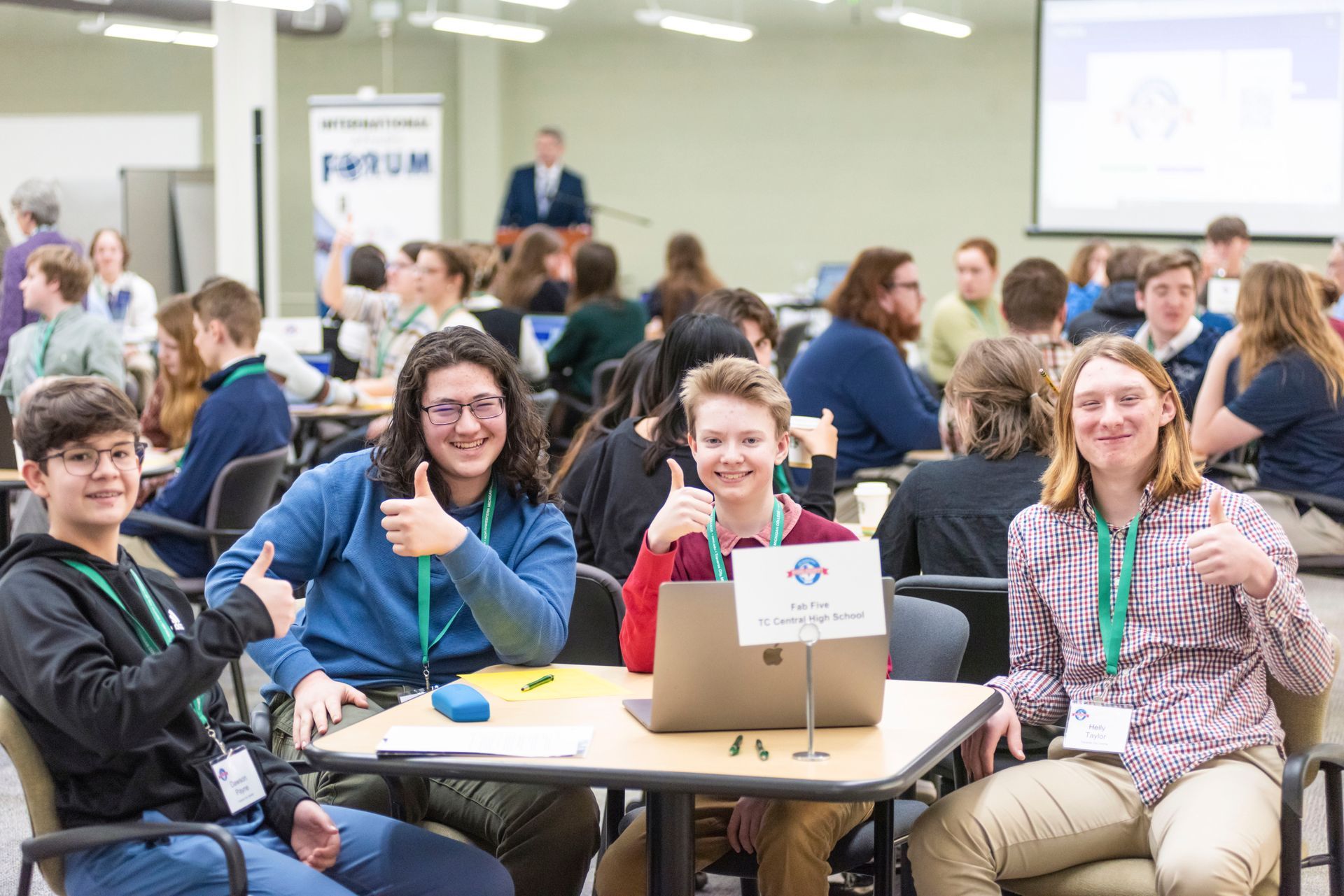 A group of young people are sitting at tables with laptops and giving thumbs up.