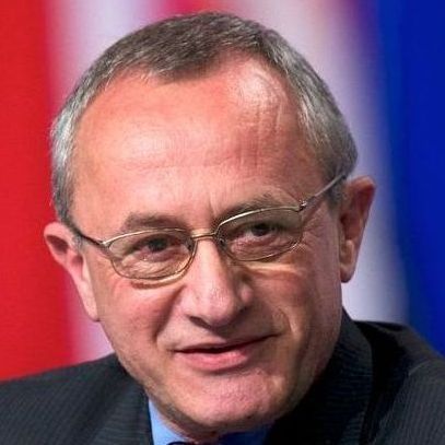 A man wearing glasses and a suit is smiling in front of a flag.