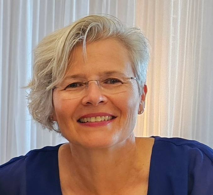 Smiling person with short silver hair and glasses, wearing a blue top against a light curtain background