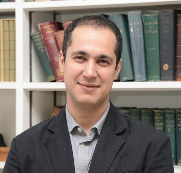 A man in a suit stands in front of a bookshelf full of books