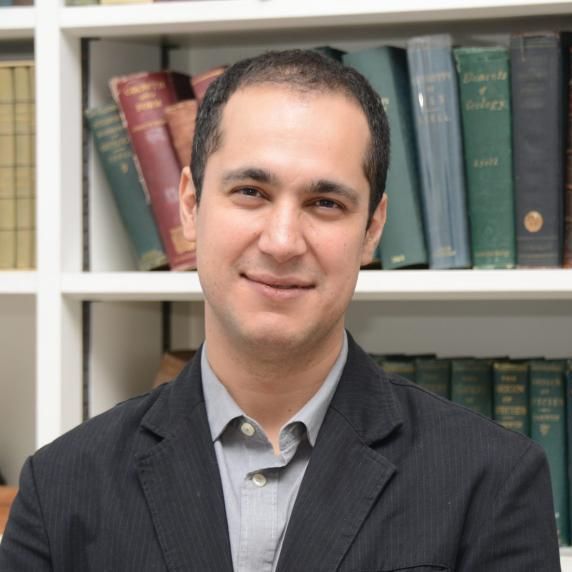 A man in a suit stands in front of a bookshelf full of books