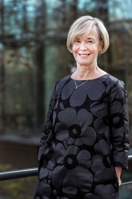 Smiling person in a dark patterned dress standing outdoors by a railing with blurred trees behind them