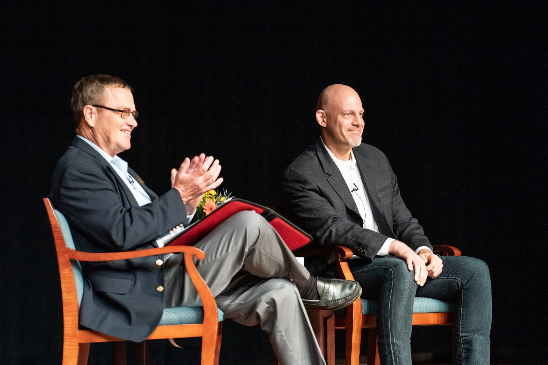 Two men are sitting in chairs on a stage and applauding.