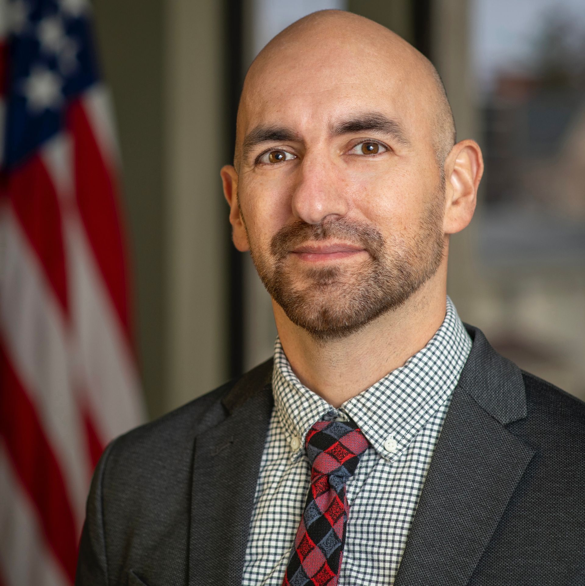 A bald man with a beard wearing a suit and tie is standing in front of an american flag.