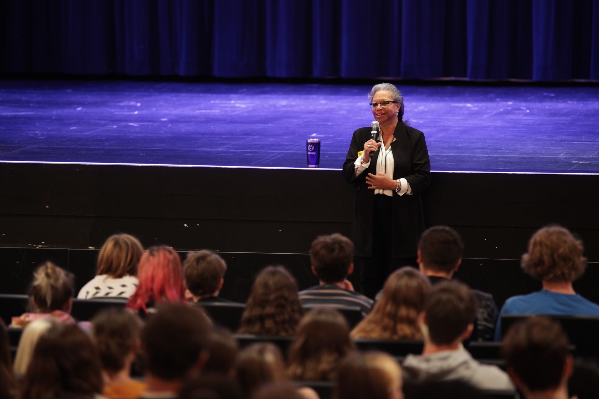 A woman is standing on a stage talking into a microphone to a crowd of people.