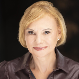 Woman with short blonde hair, wearing a brown shirt, smiles at the camera against a dark background.
