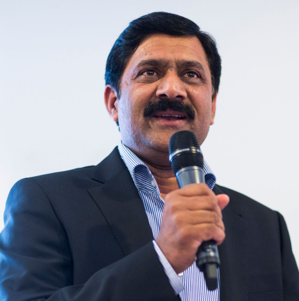Man in a suit speaks into a microphone, gesturing with his hands, in front of a white wall.
