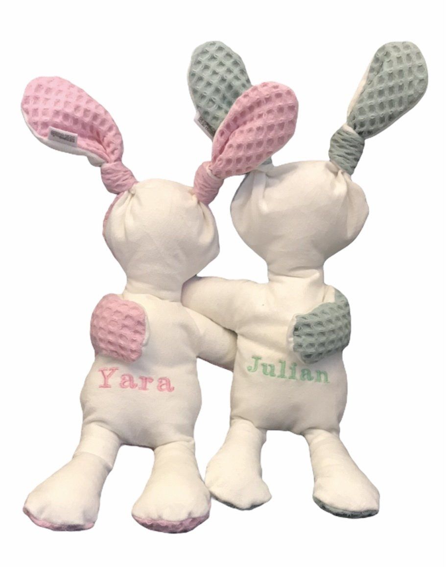 Twee witte pluche konijnen met roze en groene oren zitten naast elkaar en hebben namen op hun lijfjes geborduurd.