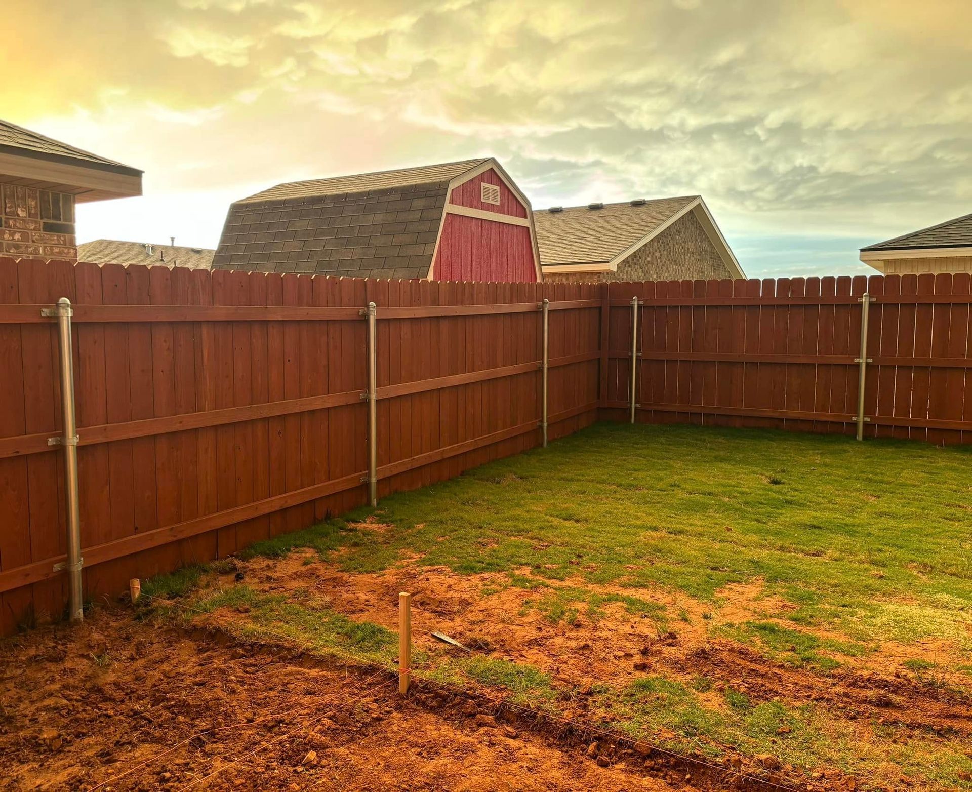Backyard with brown wooden fence, green grass, and red barn under cloudy sky.