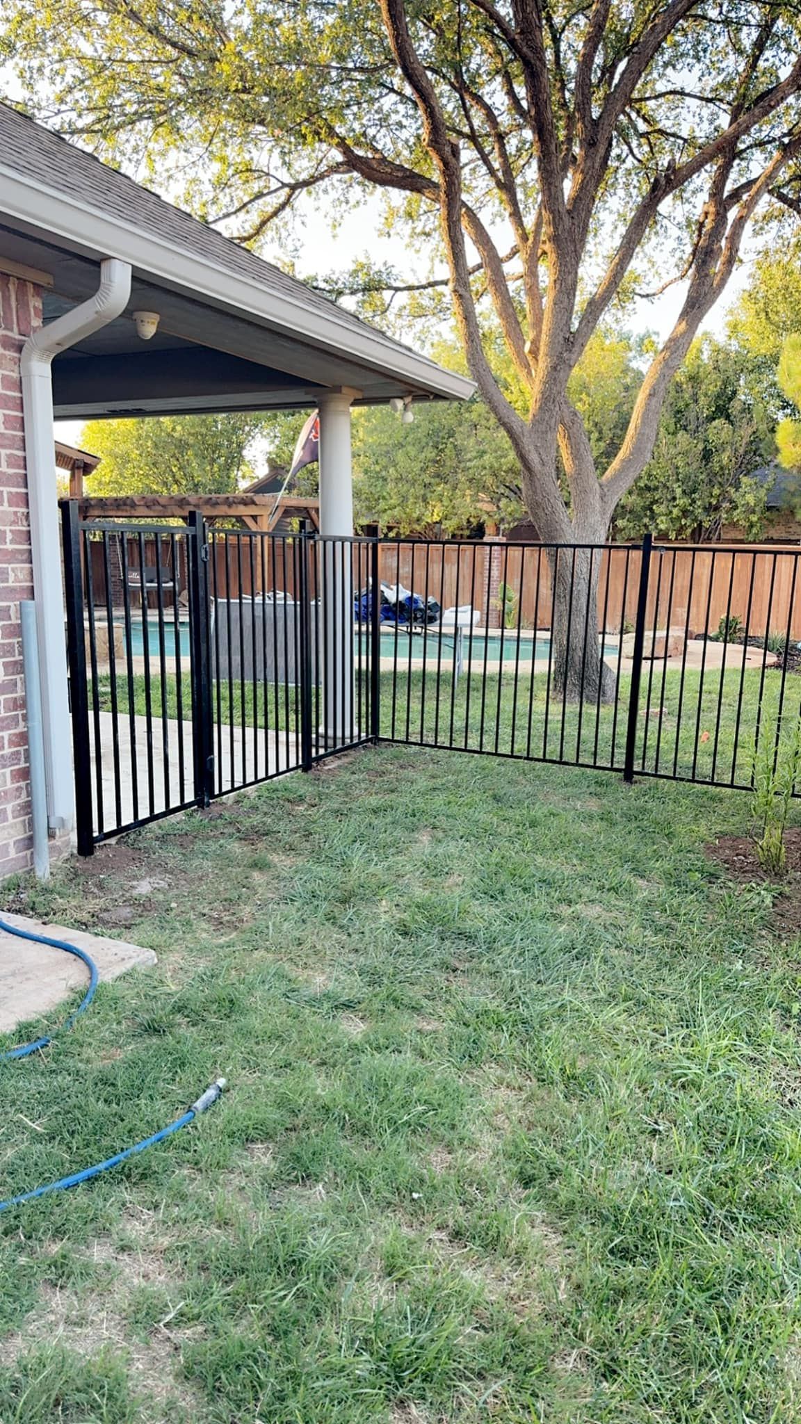Black metal fence surrounds a backyard pool, with a brick house and trees in the background.