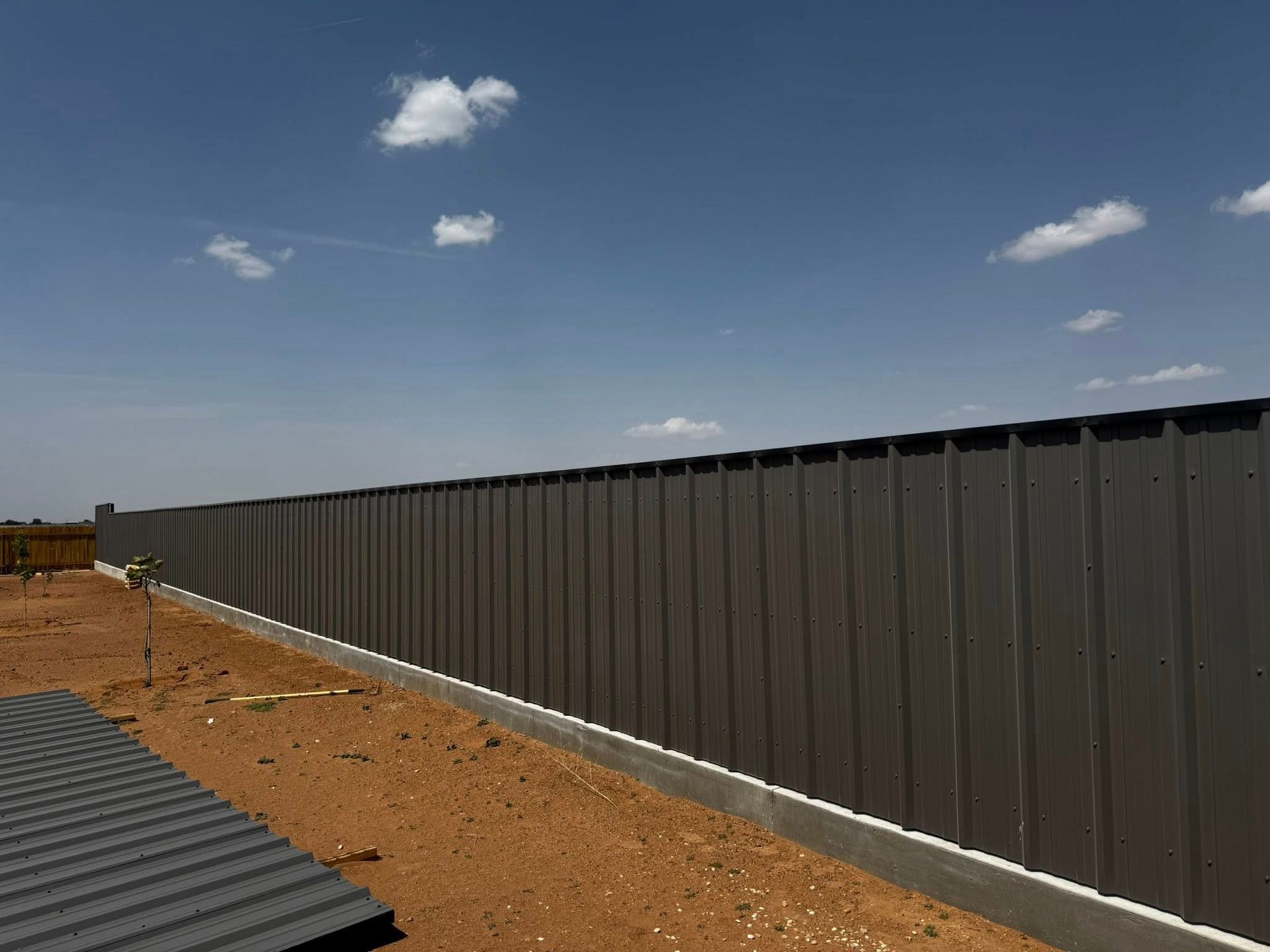Gray metal fence stretches across a barren, reddish dirt ground under a blue sky with sparse clouds.