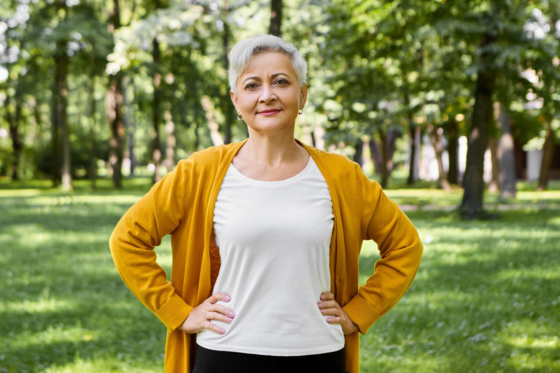 Woman with gray hair, hands on hips, smiling in a park, wearing a yellow cardigan over a white top.
