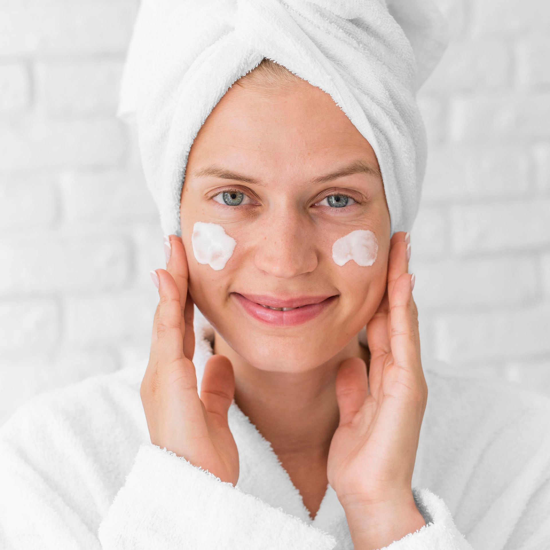 Woman in white robe and towel applying cream to cheeks, smiling.
