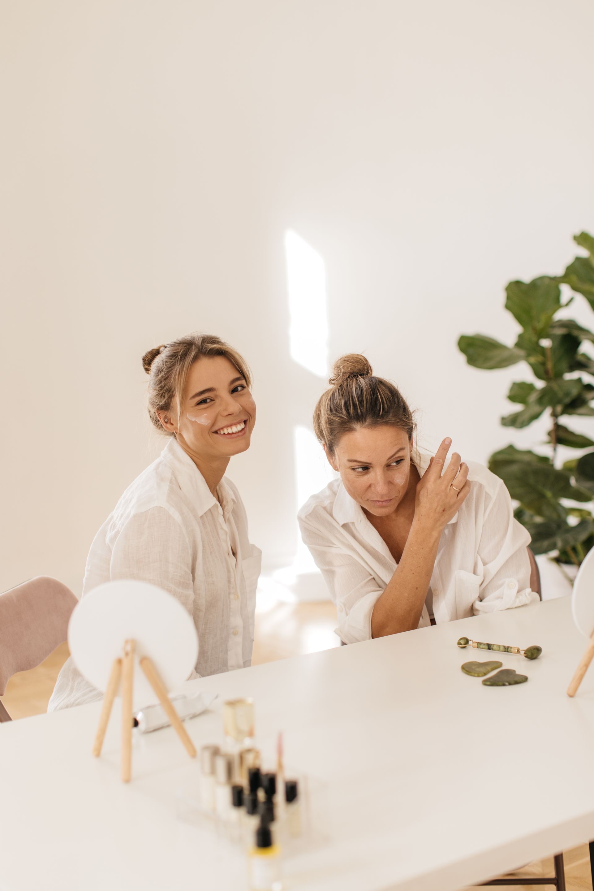 Two women in white shirts at a table, applying skincare. One smiles, the other touches her shoulder. Bright, well-lit room.