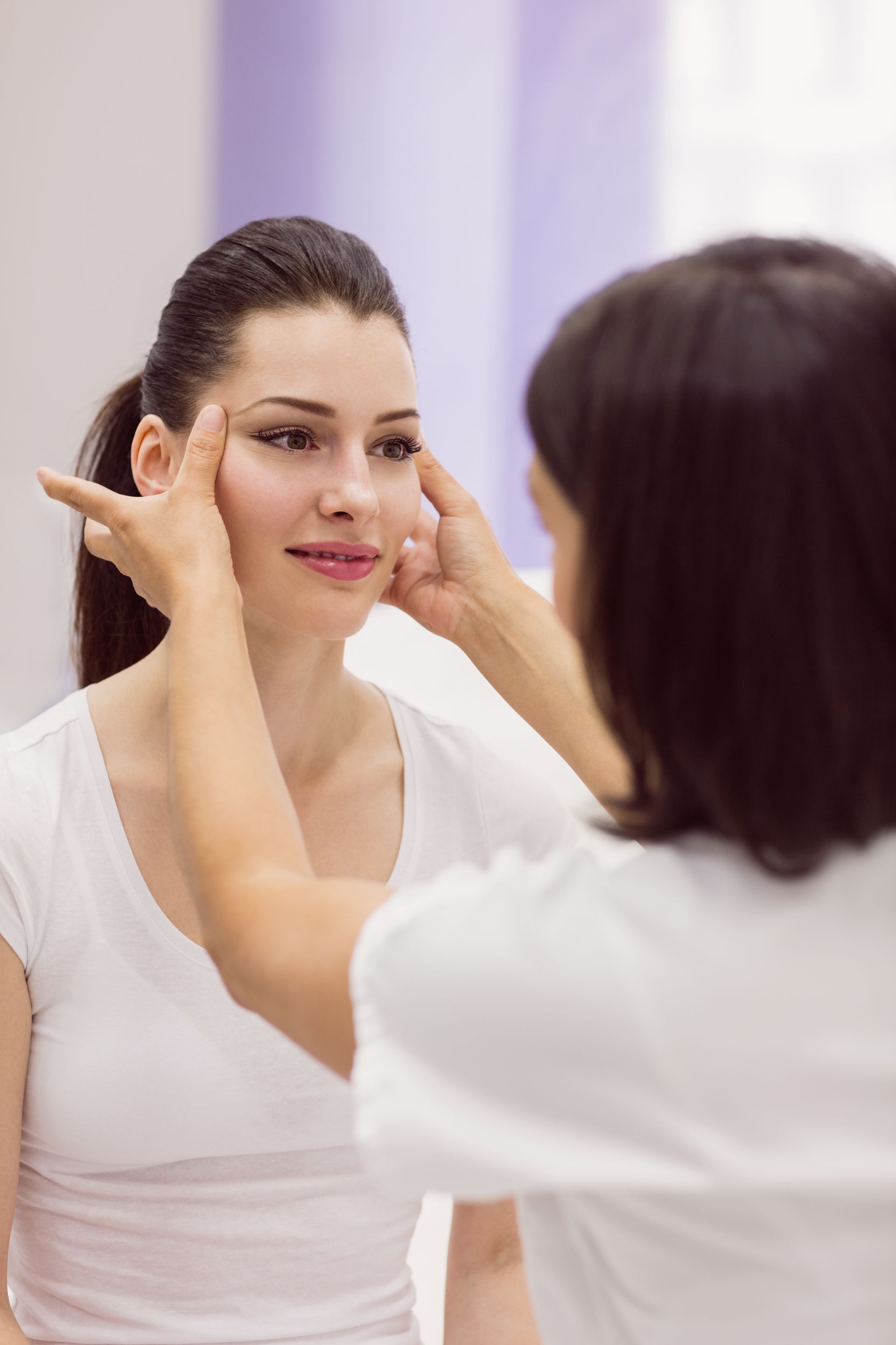 Woman's face examined by another person's hands. Both wear white shirts. Neutral background.