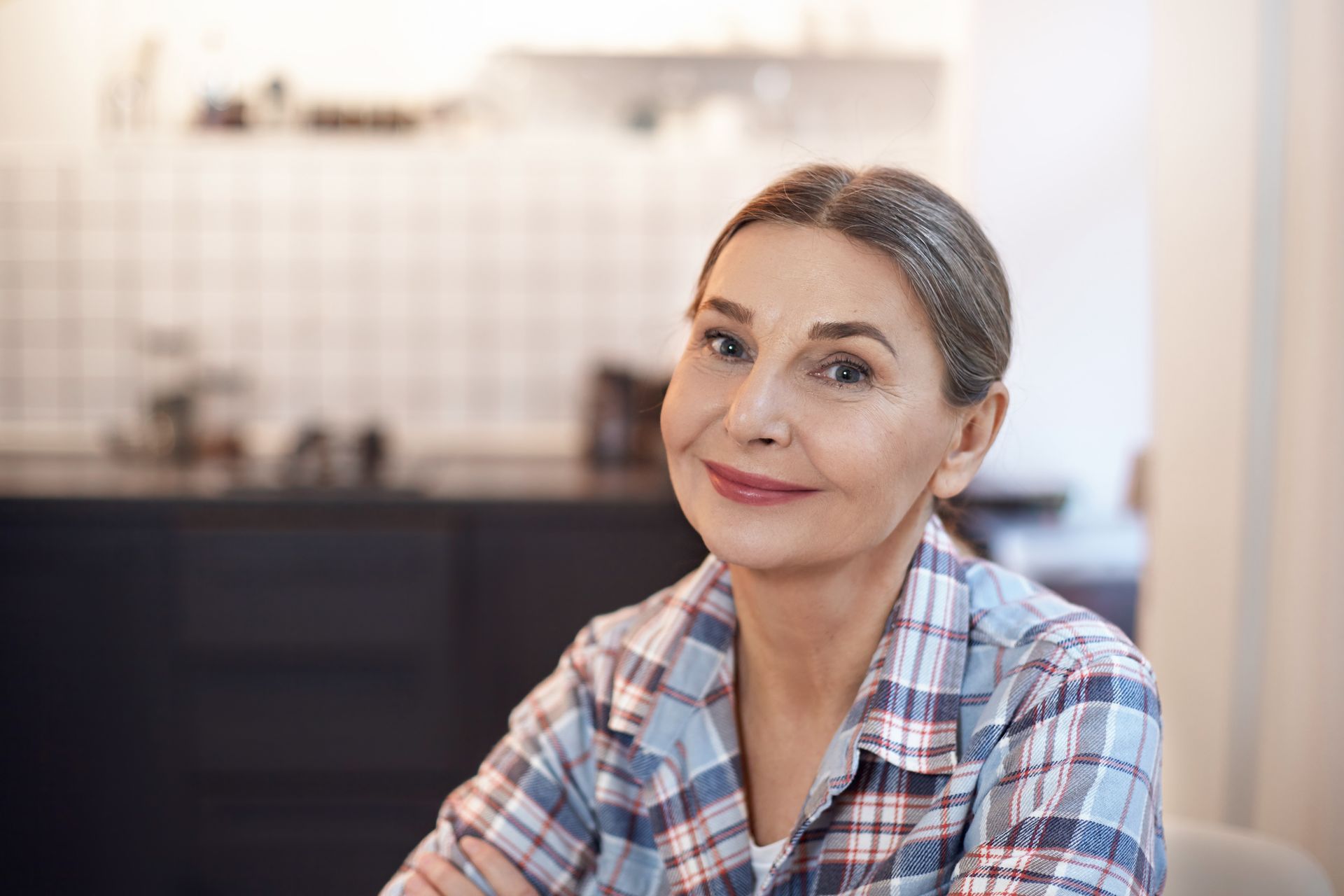 Woman in blue plaid shirt smiles indoors, kitchen background.