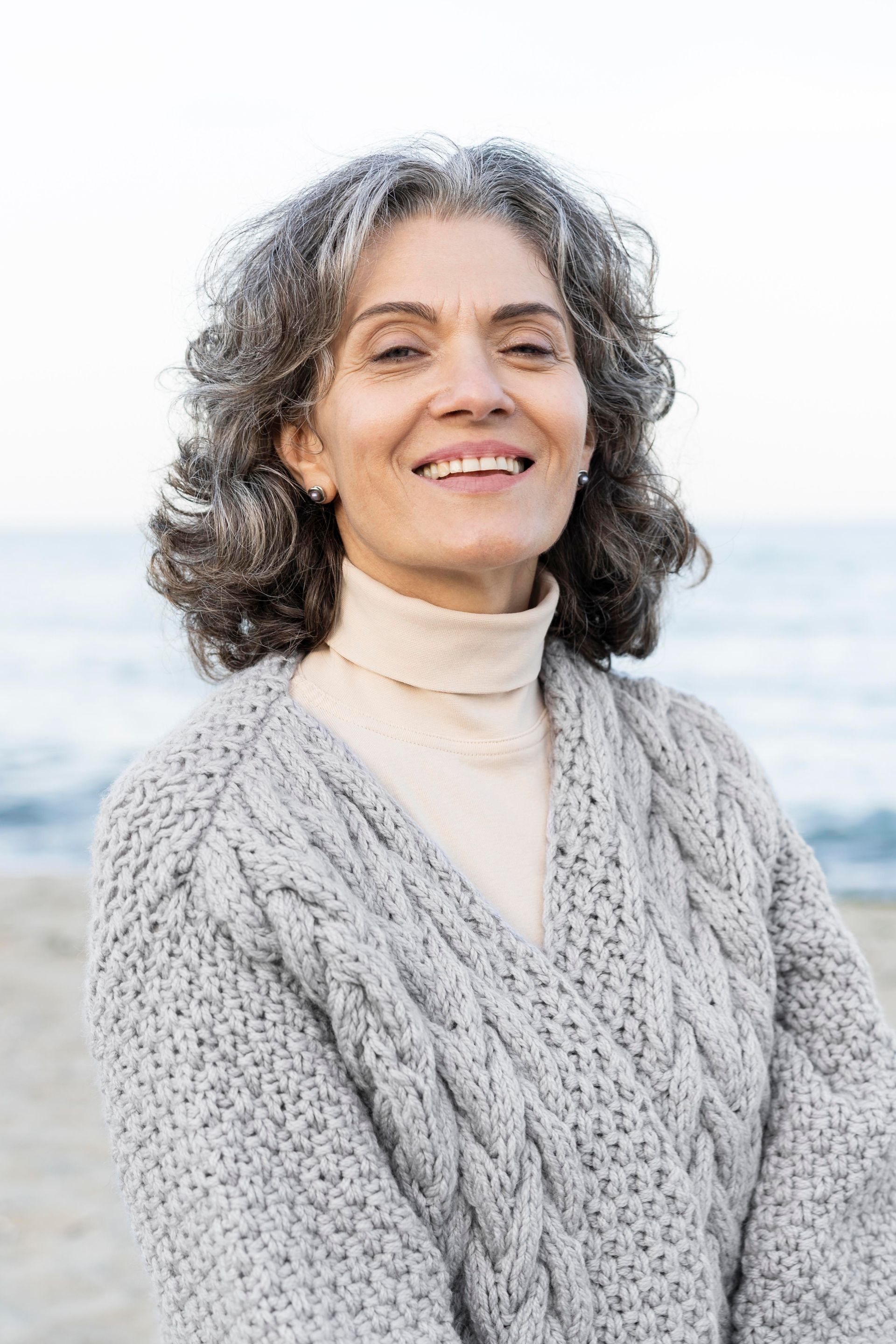 Woman with curly gray hair, smiling, wearing a cable-knit sweater and turtleneck at the beach.