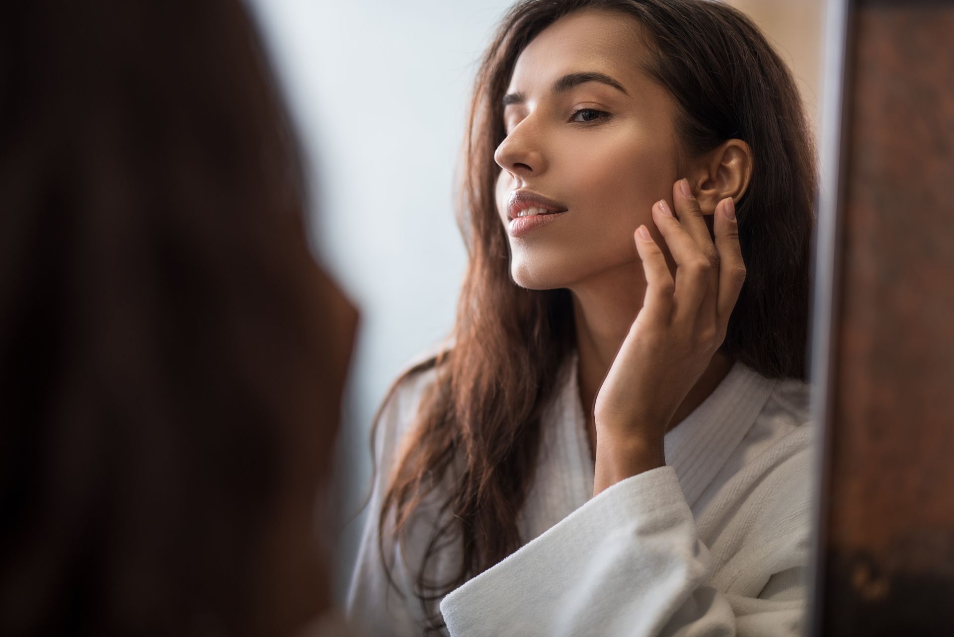 Woman in a white robe examines her face in a mirror, touching her cheek.