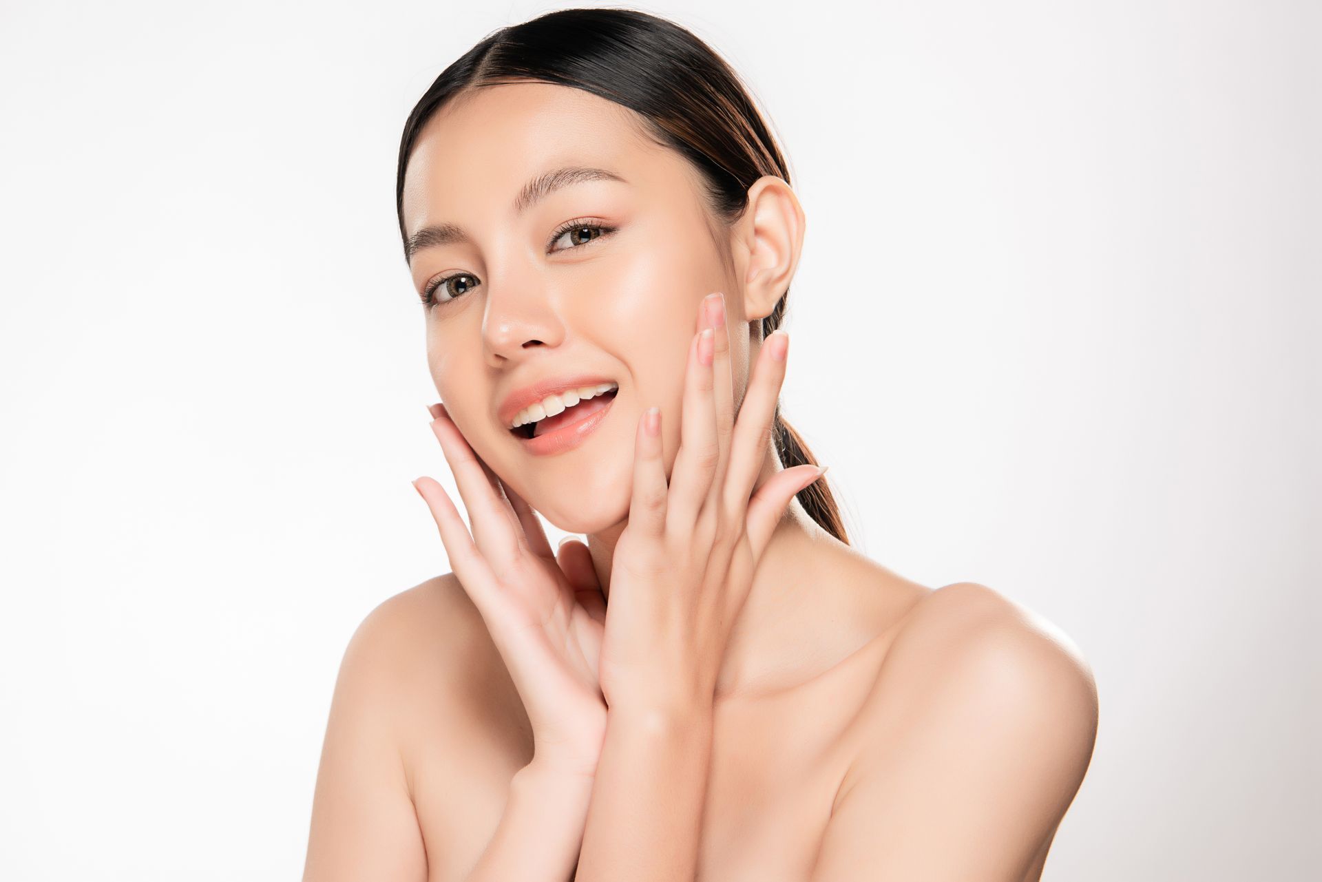 Woman with a glowing complexion smiles, hands near face, against a white background.