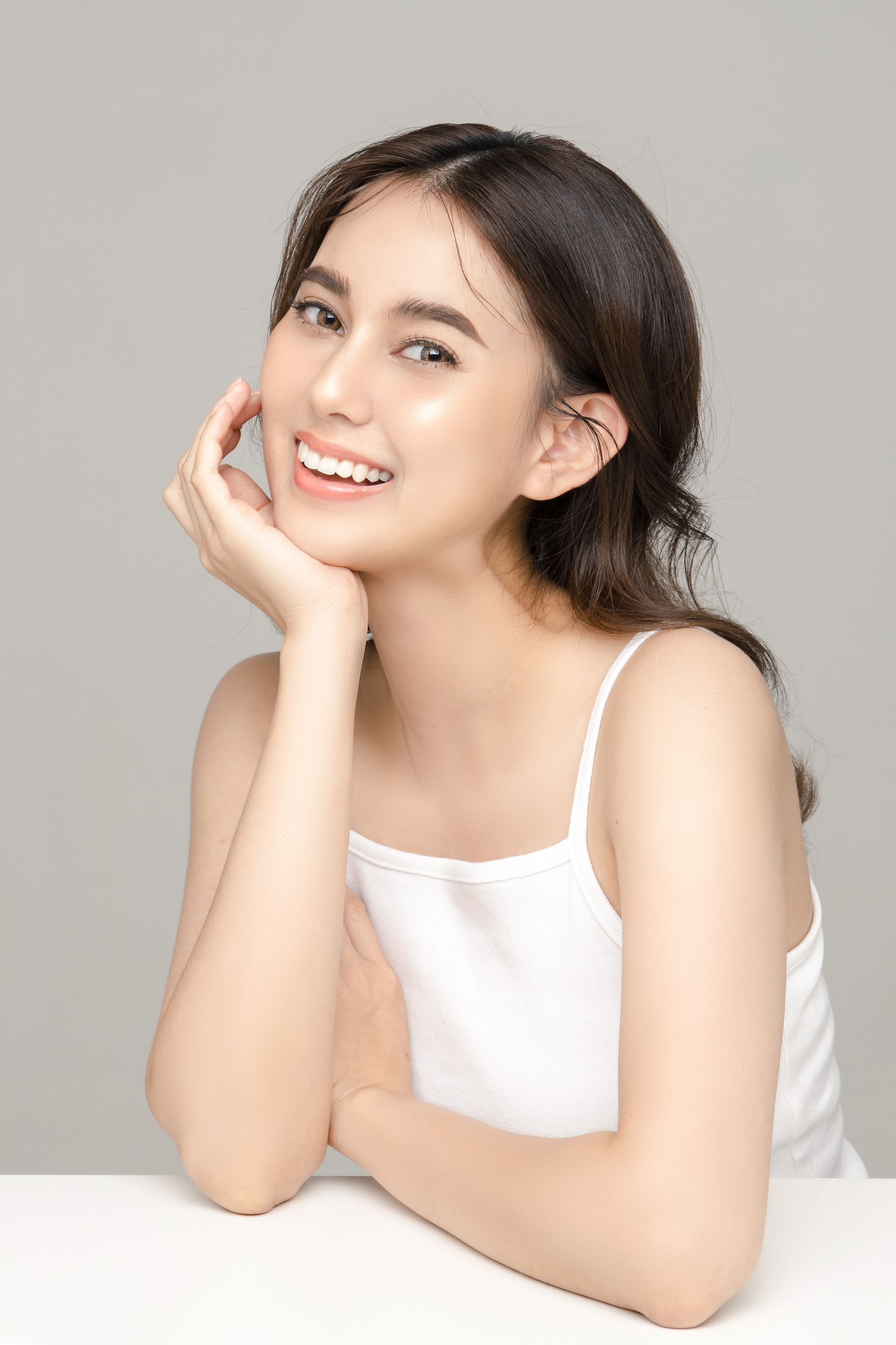 Woman smiling, resting chin on hand, wearing white tank top, against a light gray background.