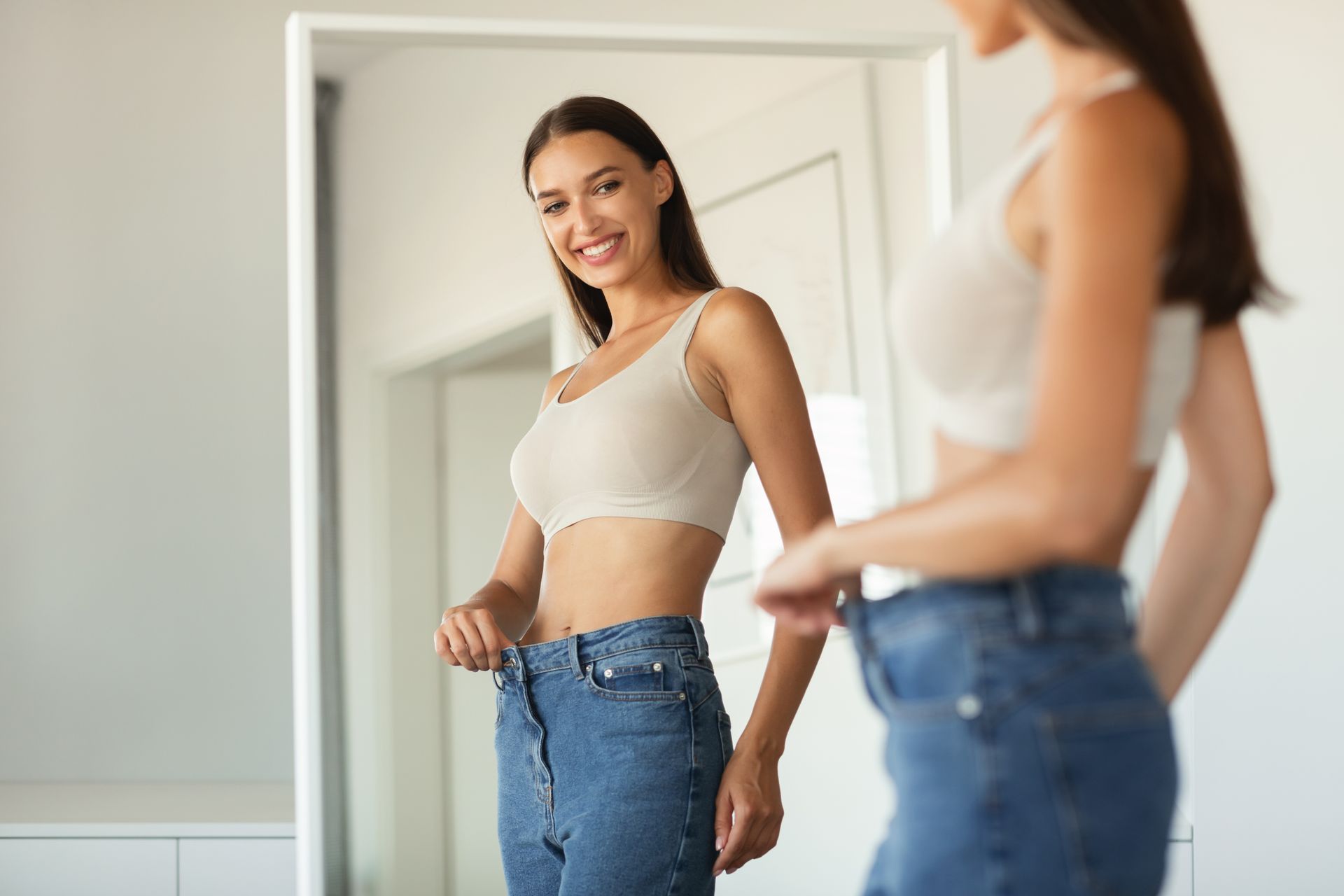 Woman smiles, holding loose jeans in front of a mirror, showing weight loss.