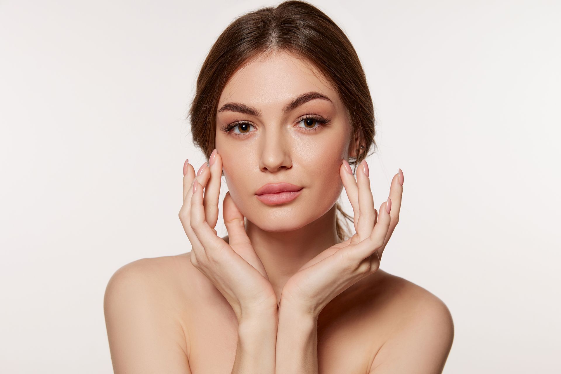 Woman with hands gently touching her cheeks, nude shoulders, soft makeup, neutral background.
