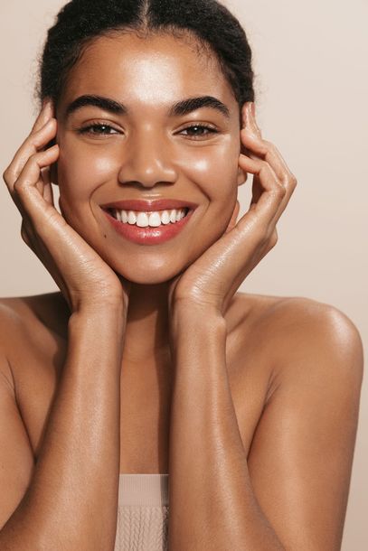 Woman with dewy skin smiles, hands framing her face, against a beige background.