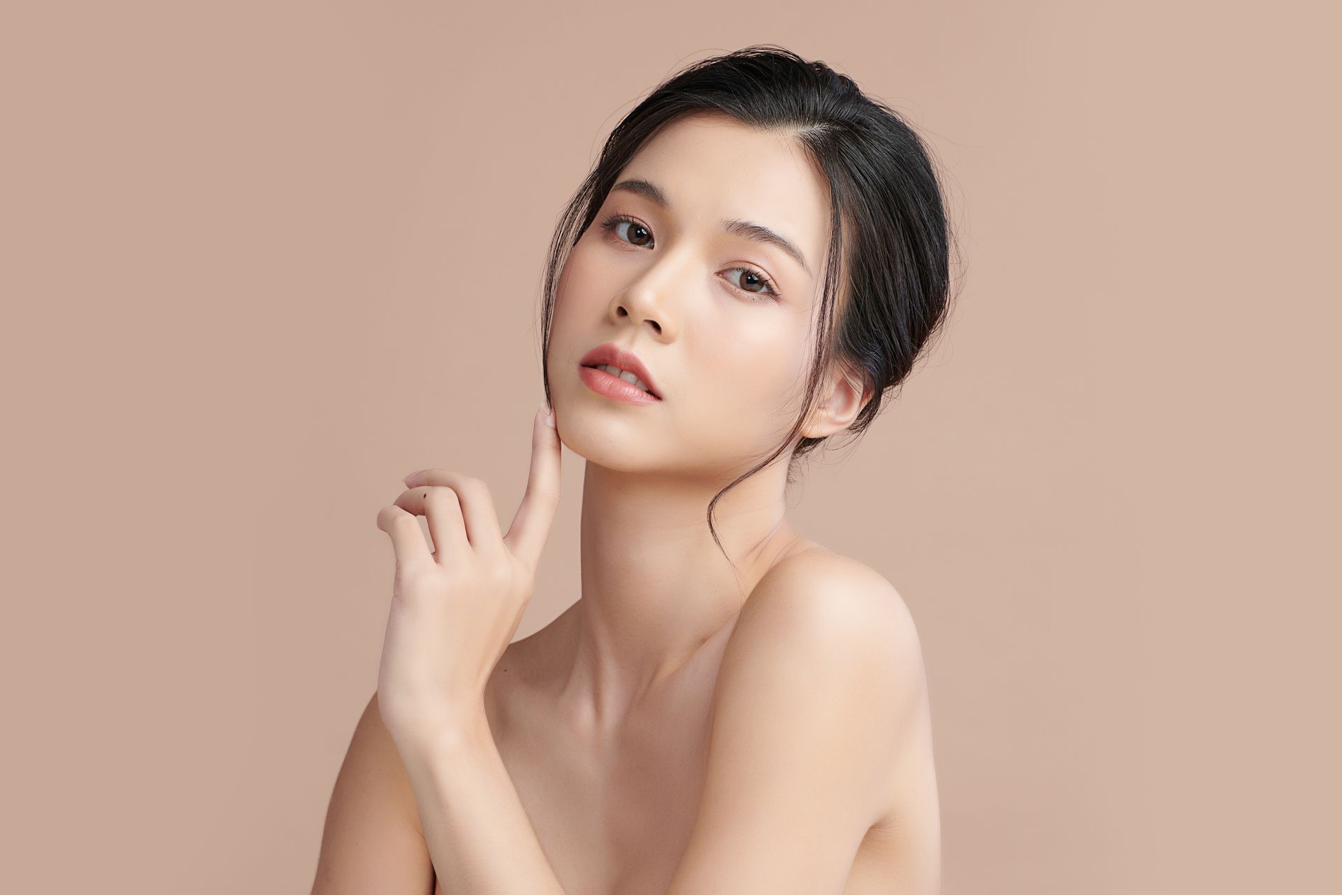 Woman with dark hair, touching her face, nude shoulders, against a light brown backdrop.