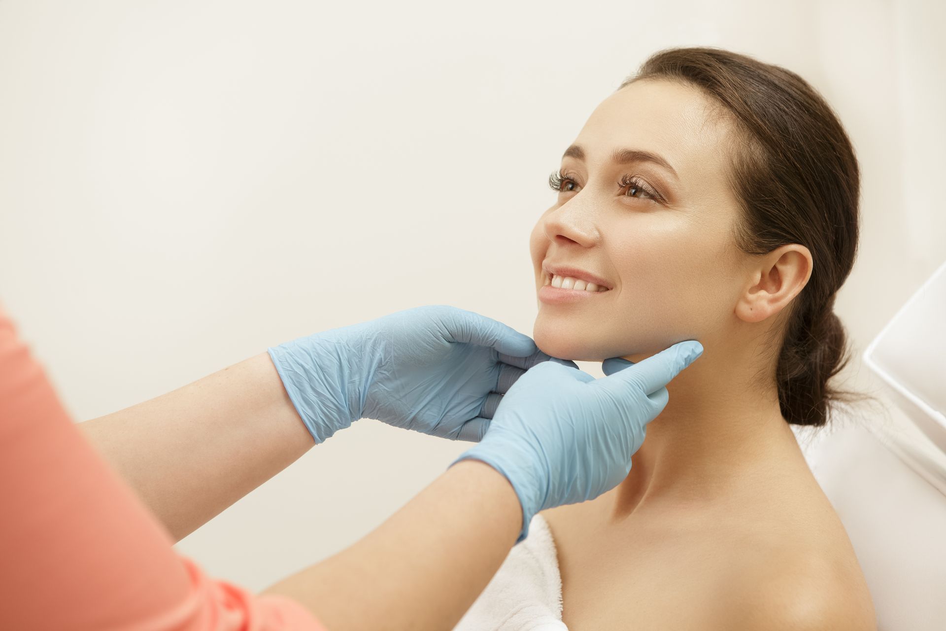 Woman smiles as hands in blue gloves examine her chin. Pale setting.
