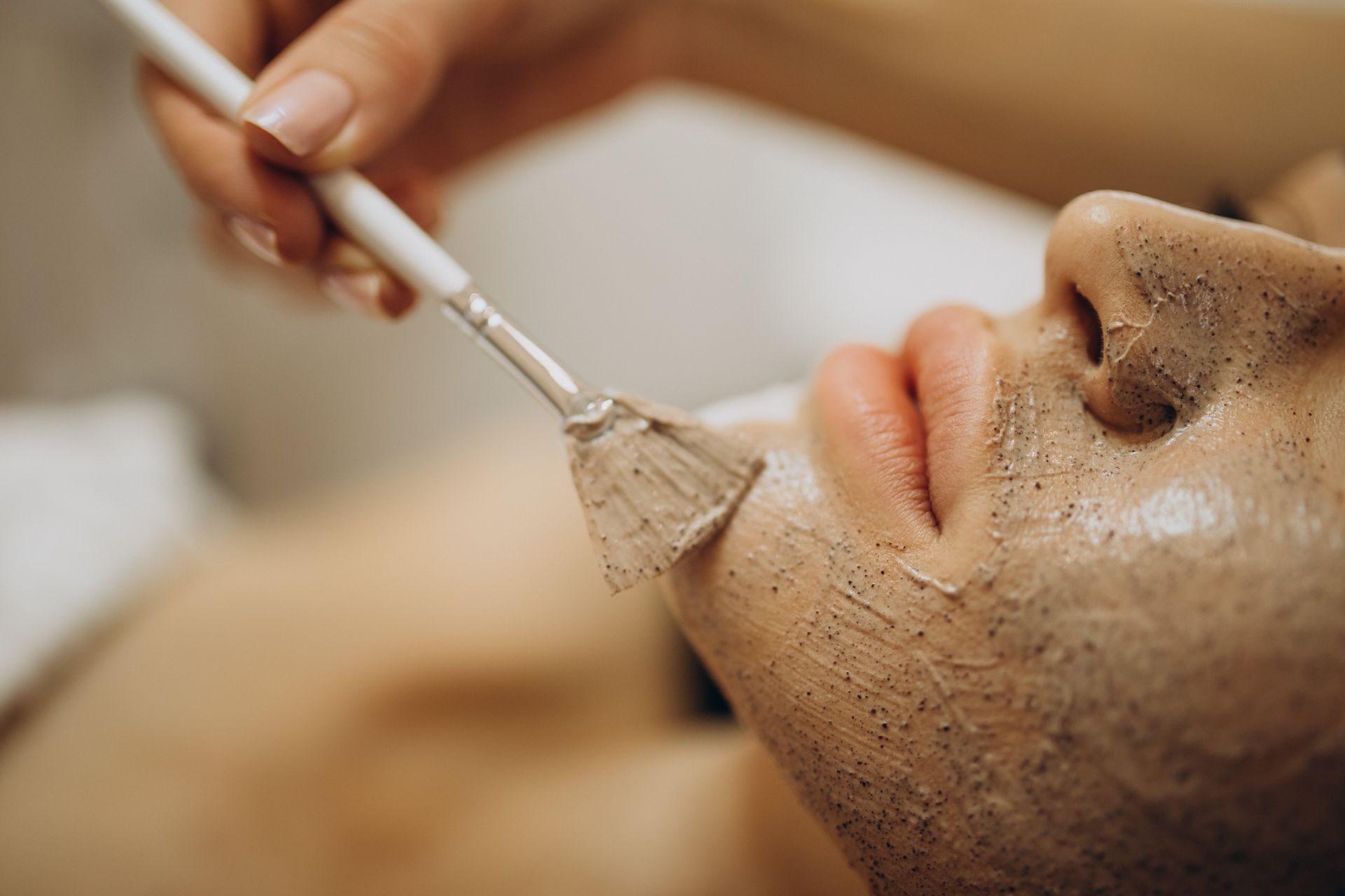 Person receiving facial treatment; cosmetician applying mask with brush.
