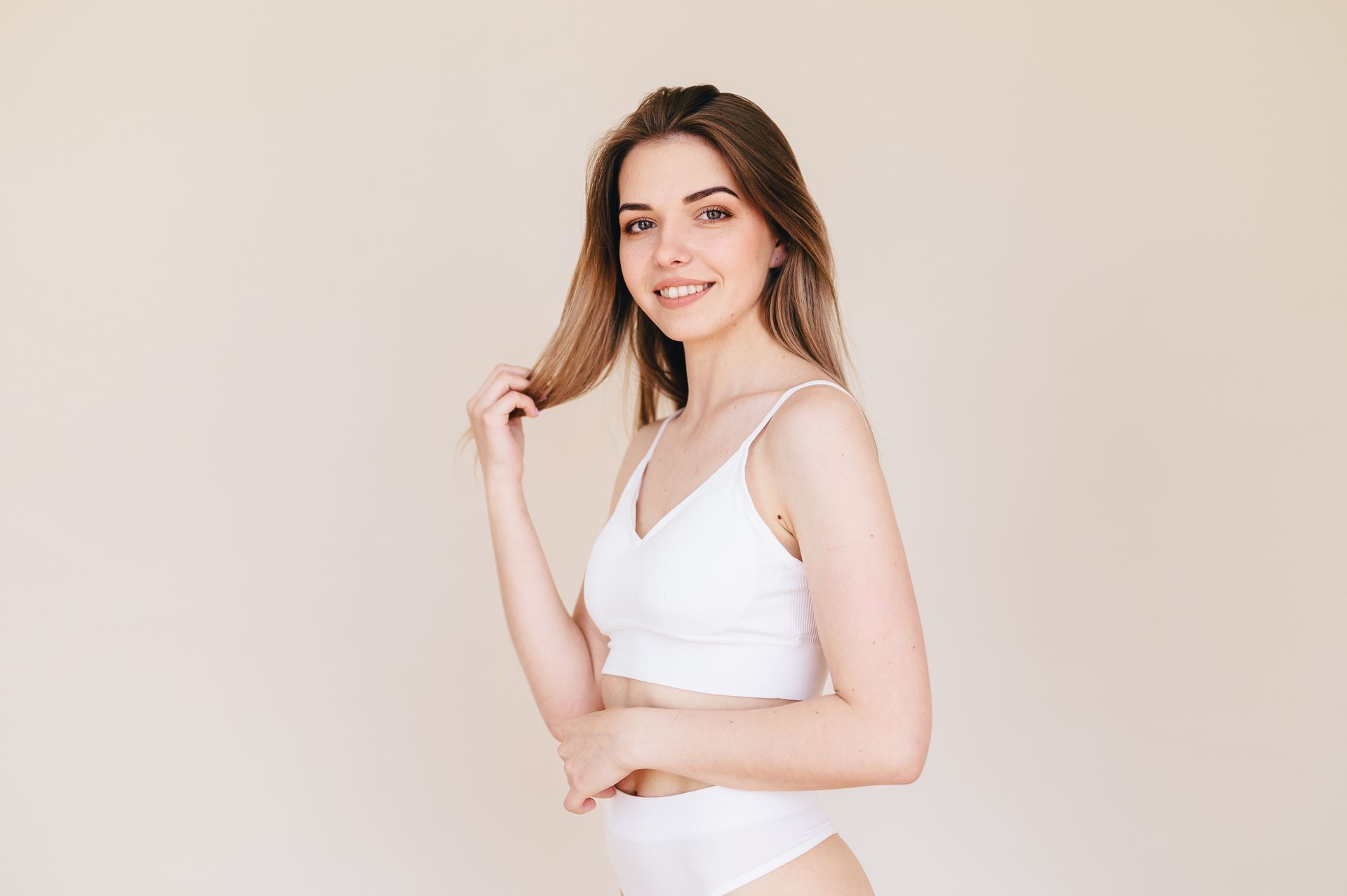 Woman in white lingerie, smiling, holding hair, against a neutral background.