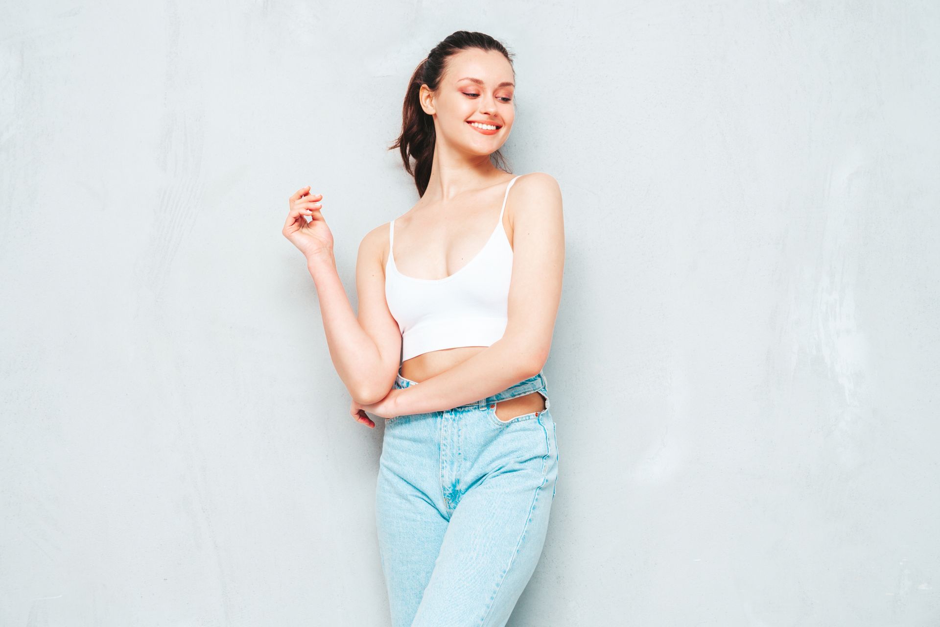 Woman wearing a white crop top and light blue jeans, smiling, posing against a gray wall.