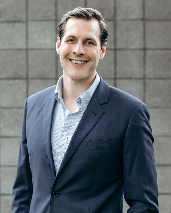 Smiling person seated against a light gray background, wearing a light blue button-up shirt and dark pants