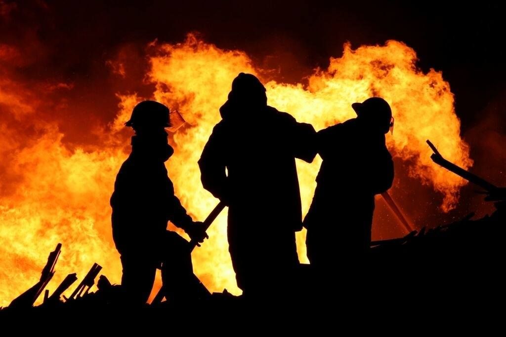 Trois personnes en silhouette devant un grand brasier, dont une tient un outil au premier plan