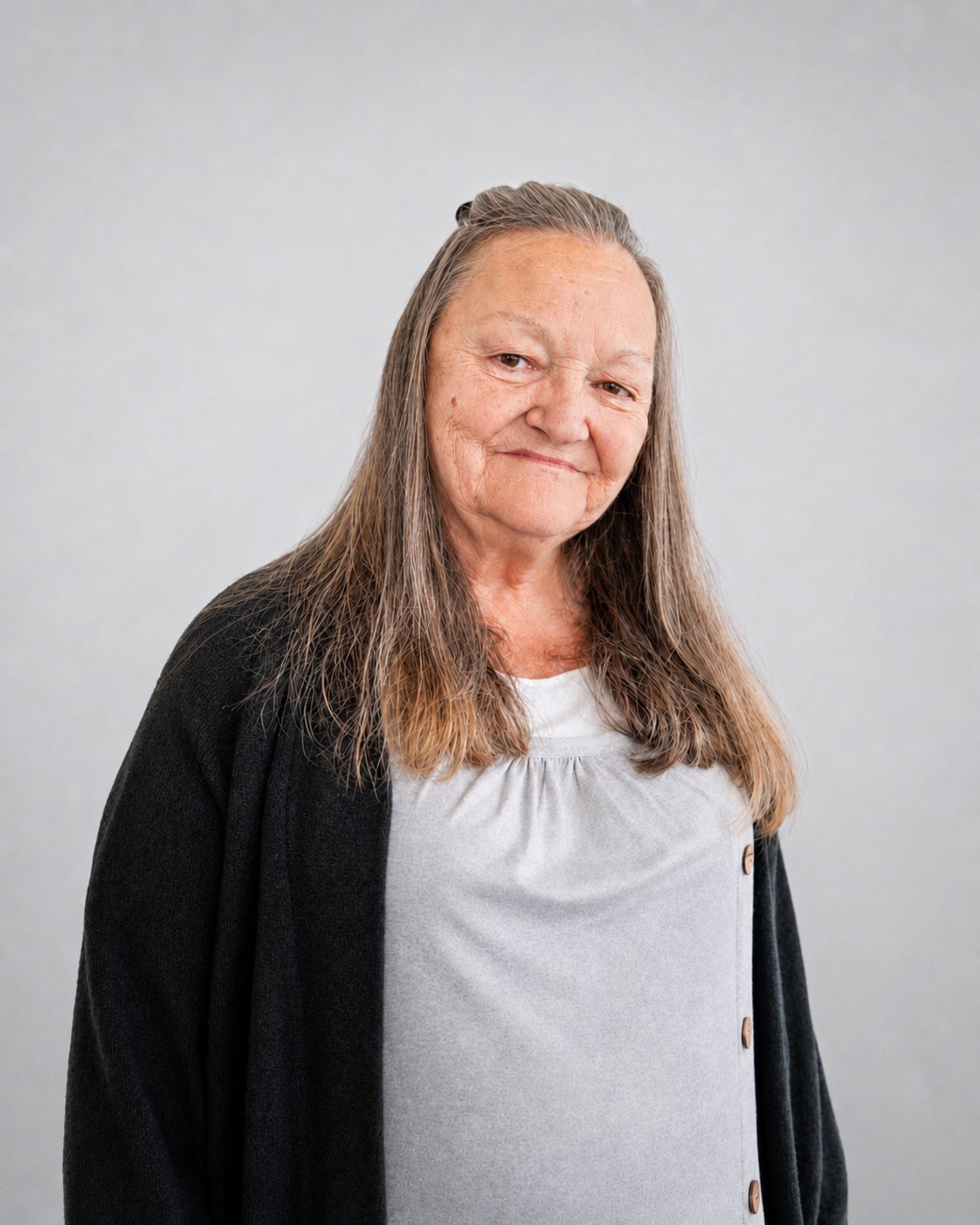 Portrait d’une personne souriante aux longs cheveux gris, portant un cardigan foncé sur un haut blanc, sur fond gris