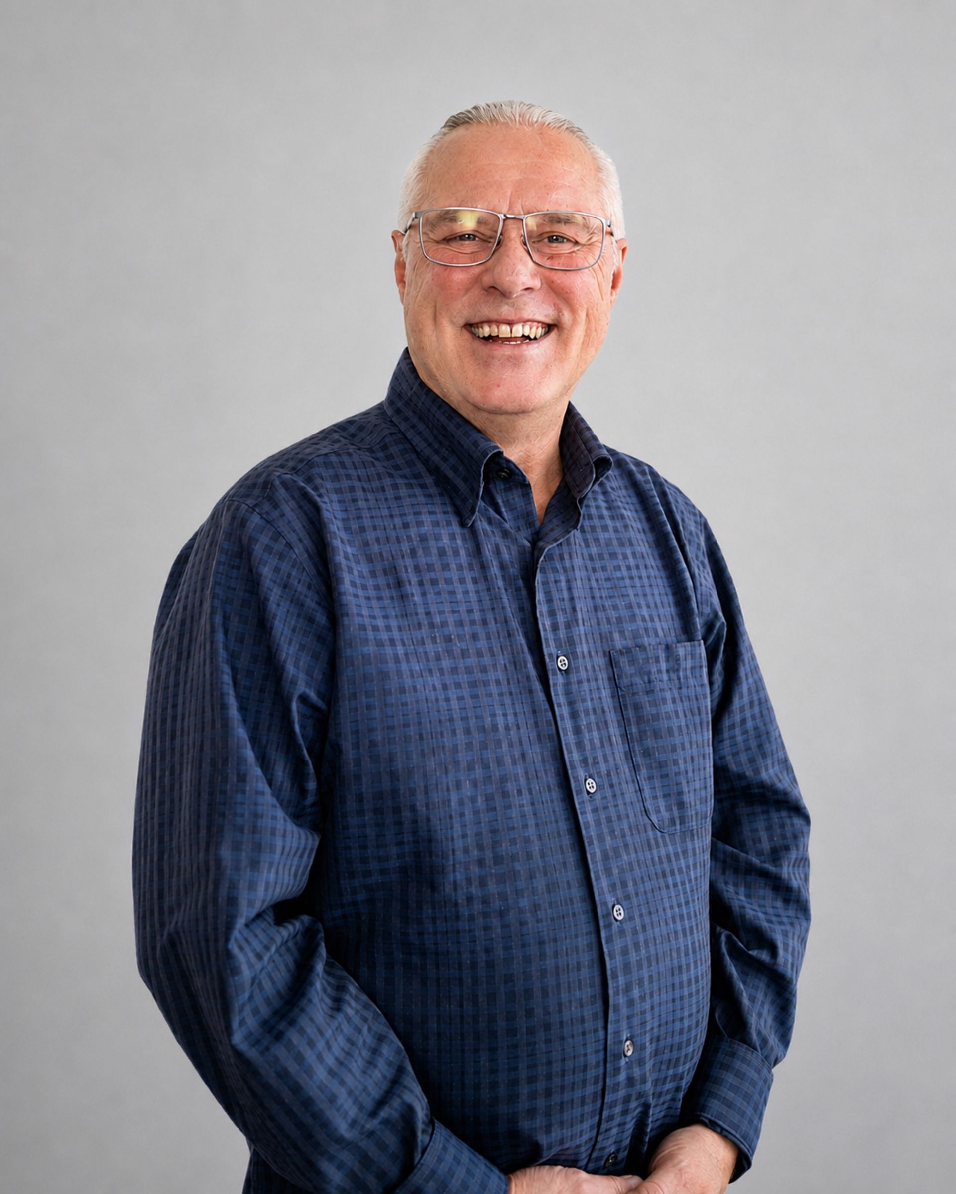 Homme souriant portant une chemise bleue boutonnée et des lunettes, debout devant un fond gris uni.