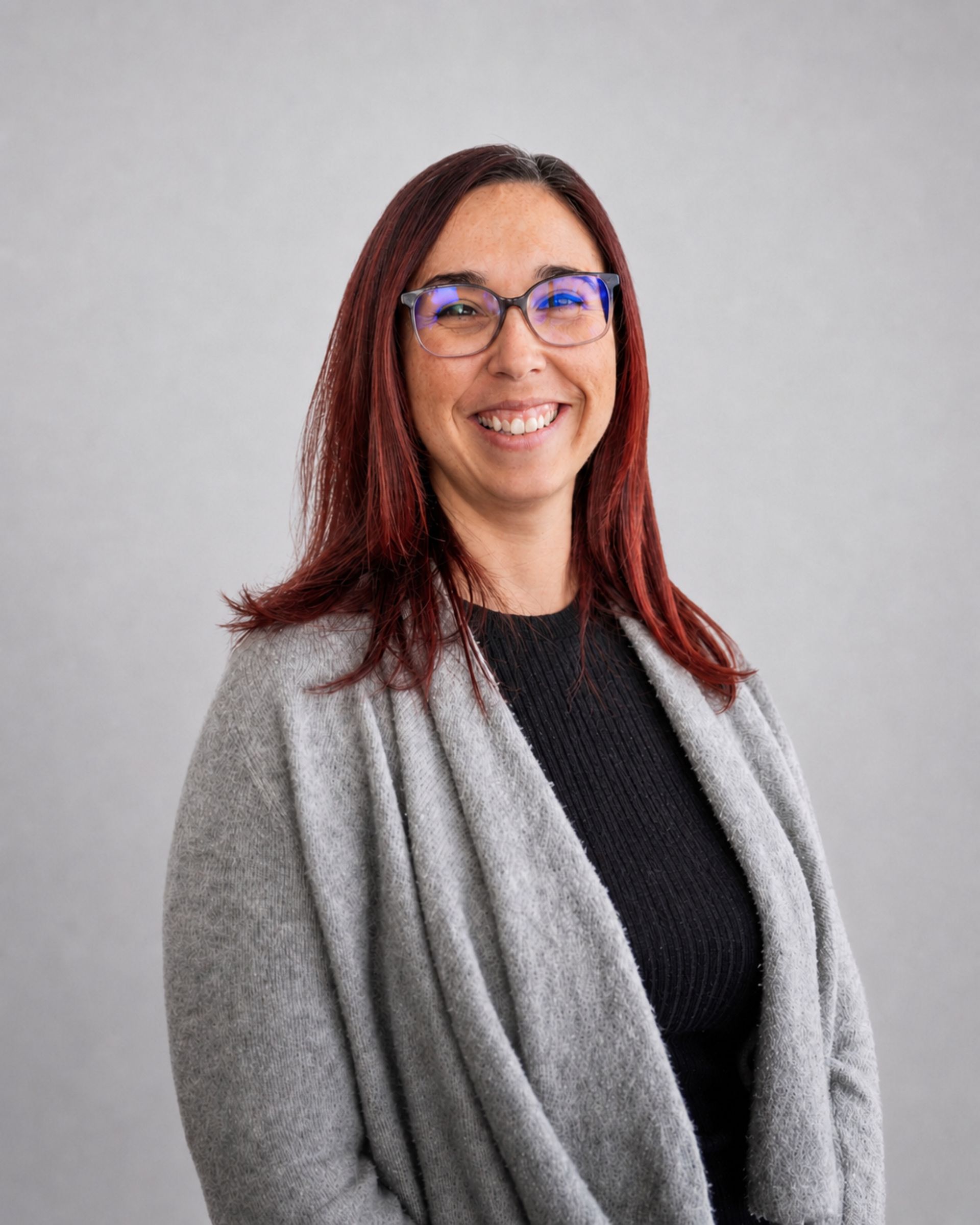 Personne souriante portant des lunettes et un châle gris sur un haut noir, sur fond gris clair