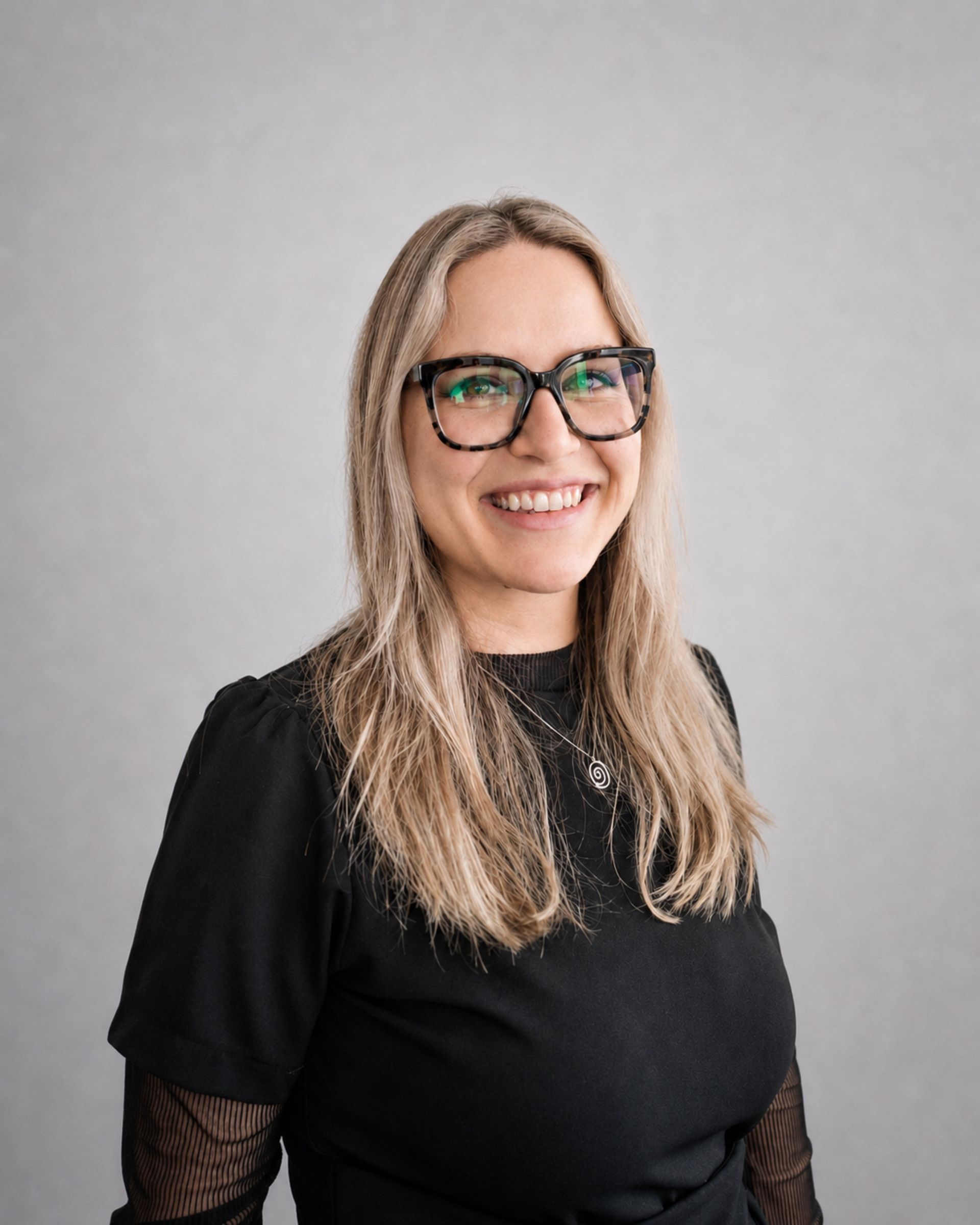 Femme souriante portant des lunettes et les cheveux longs blonds, vêtue d’un haut noir sur fond gris