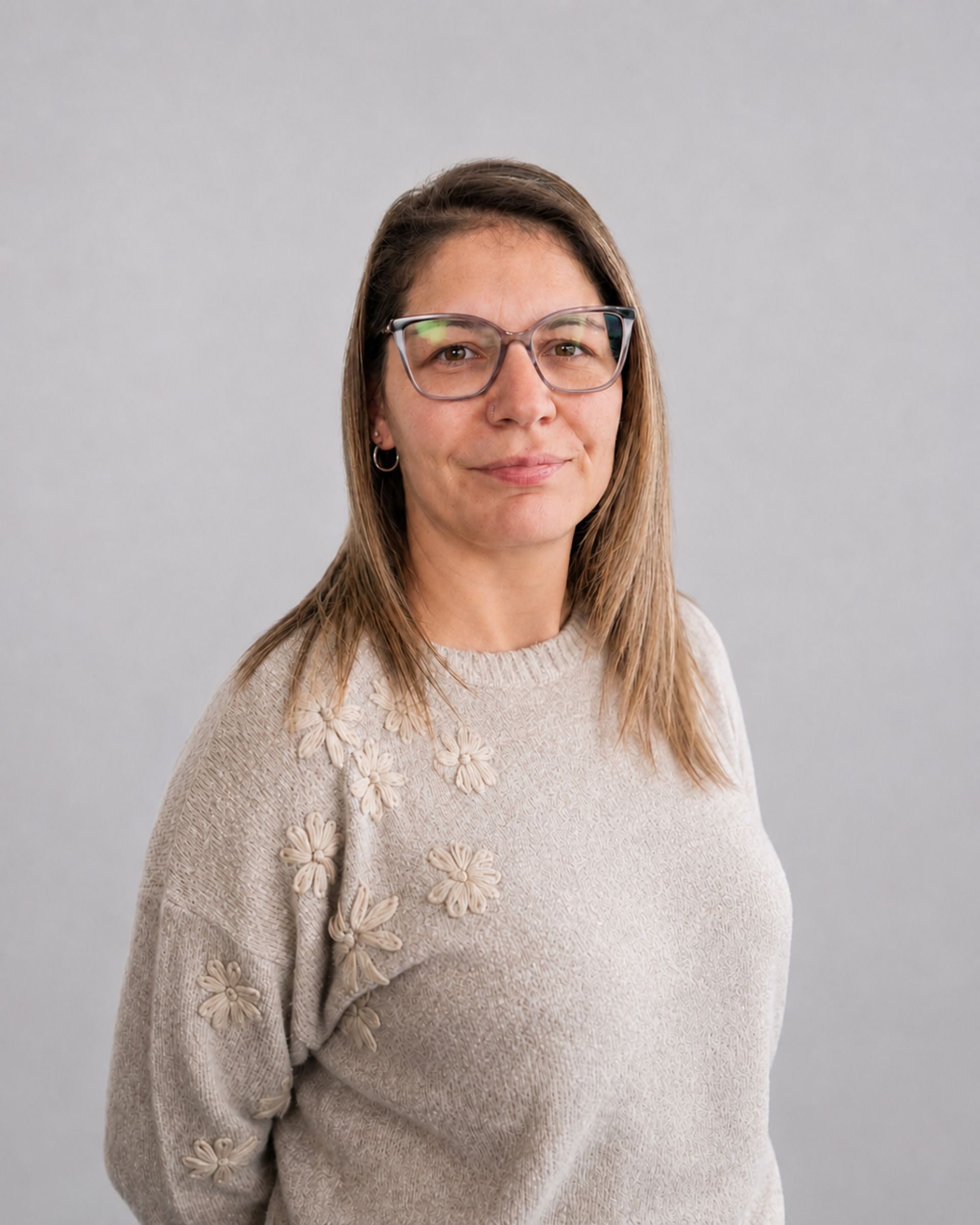 Femme portant des lunettes et un pull beige, debout devant un fond gris uni.