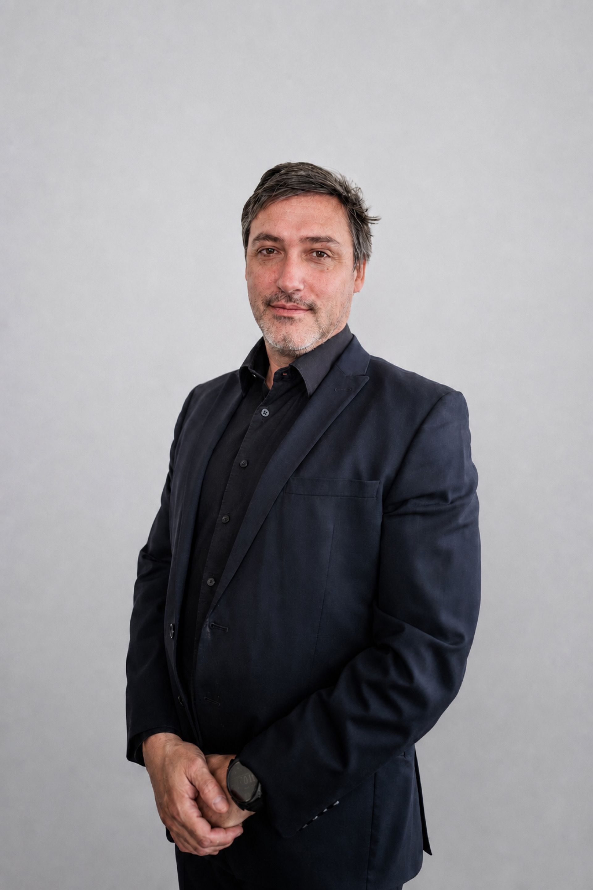 Une personne en costume sombre se tient devant un fond gris clair, face à l’appareil photo.